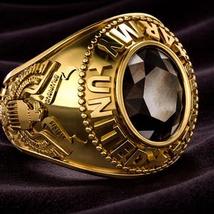 May include: A gold-colored ring with a large, dark gemstone at its center. The ring features intricate detailing, including the words "UNITED ARMY" and other inscriptions. The ring is set against a dark, textured background.
