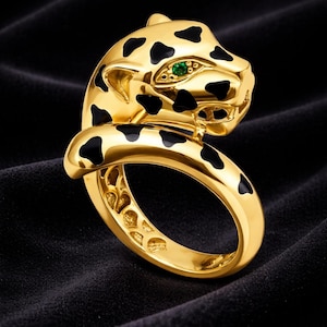 Könnte beinhalten: Ein goldfarbener Ring mit einem stilisierten Leopardenkopf, schwarzen Flecken und einem grünen Edelsteinauge. Der Ring hat eine glatte, polierte Oberfläche und ist vor dunklem Hintergrund platziert.