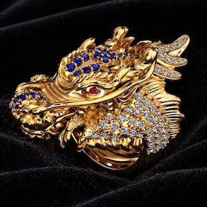 Könnte beinhalten: Ein goldener Drachenring mit detaillierten Verzierungen. Der Drachenkopf ist mit blauen und klaren Edelsteinen sowie einem roten Edelsteinauge besetzt. Der Ring liegt auf schwarzem Hintergrund.
