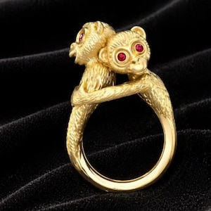 Puede incluir: Un anillo dorado con dos figuras de monos abrazándose. Los monos tienen ojos de piedras preciosas rojas. El anillo está sobre un fondo de terciopelo negro, resaltando los detalles del diseño.