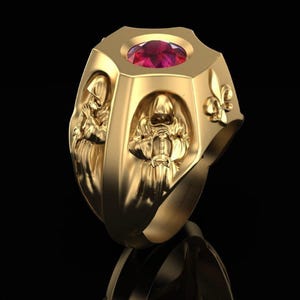 Könnte beinhalten: Ein goldfarbener Ring mit einem großen, ovalen, roten Edelstein oben. Der Ring weist detaillierte Reliefs von Kapuzenfiguren auf, die Schwerter halten. Auf der Seite befindet sich ein Fleur-de-Lis-Design. Der Ring wird vor schwarzem Hintergrund präsentiert.