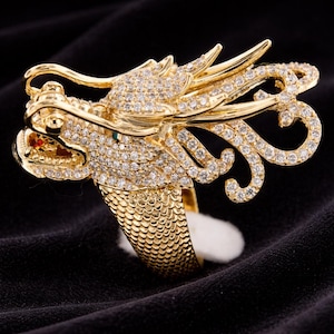 Könnte beinhalten: Ein goldfarbener Drachenring mit aufwendigen Details. Der Drachenkopf ist mit kleinen, funkelnden Steinen besetzt, und der Ring hat ein strukturiertes Band. Das Auge des Drachen ist grün, und der Mund hat einen roten Stein. Der Ring wird auf schwarzem Hintergrund präsentiert.