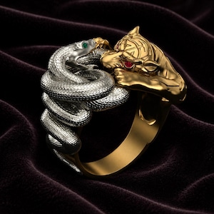 Könnte beinhalten: Ein zweifarbiger Ring mit einer silberfarbenen, gewundenen Schlange mit einem grünen Edelsteinauge und einem goldfarbenen Tigerkopf mit roten Edelsteinaugen. Schlange und Tiger stehen sich gegenüber und bilden die Ringschiene aus Metall.