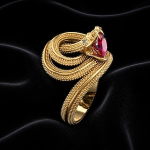 Könnte beinhalten: Ein goldfarbener Ring in Form einer aufgerollten Schlange. Der Schlangenkopf hat einen großen, oval geschliffenen roten Edelstein und einen kleinen Diamanten. Der Ring ist vor einem schwarzen Hintergrund platziert.