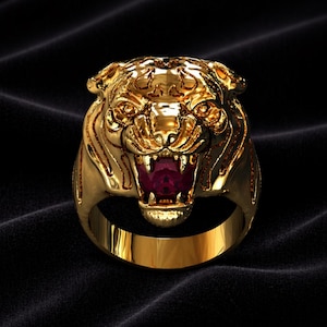 Könnte beinhalten: Ein goldfarbener Ring mit einem detaillierten Tigerkopf-Design. Das Maul des Tigers ist geöffnet und zeigt einen roten Edelstein. Der Ring ist vor einem dunklen, strukturierten Hintergrund platziert, der die Details hervorhebt.