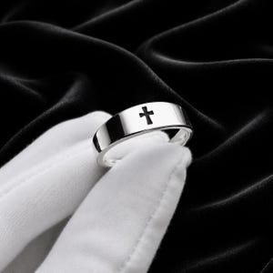 Könnte beinhalten: Ein silberner Ring mit einem Kreuz-Design. Der Ring wird auf einem weißen Handschuh vor schwarzem Hintergrund gezeigt. Das Kreuz ist eine einfache, solide Form, und der Ring hat eine polierte Oberfläche.