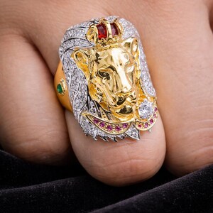 May include: A gold-toned lion head ring with a red gem crown and diamond accents. The lion's mane is detailed with white stones, and the ring features a green and pink gem. This statement ring is a bold piece of jewelry.