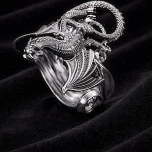 Könnte beinhalten: Ein silberner Drachenring mit detaillierten Merkmalen. Der Drache umschlingt das Band, wobei Flügel und Schuppen sichtbar sind. Der Ring ist vor einem schwarzen Hintergrund platziert, der den metallischen Glanz hervorhebt.