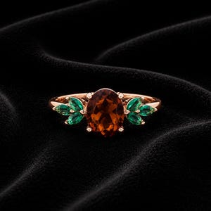 Könnte beinhalten: Roségoldener Ring mit einem ovalen, rotbraunen Edelstein, flankiert von grünen, blattförmigen Akzenten. Der Ring ist vor einem dunklen, strukturierten Hintergrund platziert, der das Design und den Farbkontrast hervorhebt.
