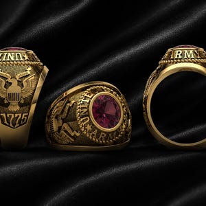 Könnte beinhalten: Ein goldfarbener Ring mit einem runden, roten Edelstein in der Mitte. Das Band ist mit dem Text UNITED STATES ARMY und dem Jahr 1775 graviert. Die Seite zeigt ein Adleremblem mit einem Banner mit der Aufschrift COUNTRY HONOR und Seil-Details.