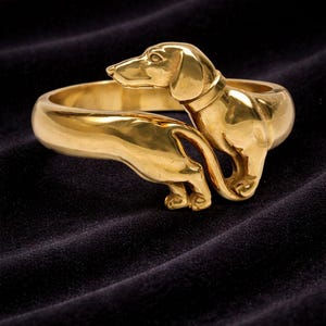 Könnte beinhalten: Ein goldfarbenes Dackel-Armband. Das Armband zeigt eine detaillierte Skulptur eines Dackels, dessen Kopf und Schwanz die Enden des Armbands bilden. Der Hund trägt ein Halsband. Das Armband besteht aus glänzendem Metall.