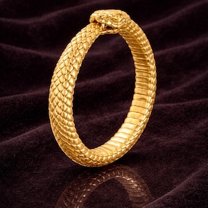 May include: A gold snake bracelet. The bracelet is in the shape of a snake biting its own tail, with detailed scales and a snake head. The jewellery is displayed on a dark, textured surface.