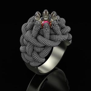 Può includere: Un anello in metallo scuro caratterizzato da un gruppo di serpenti intrecciati con texture complessa e squame dettagliate. I serpenti sono disposti in formazione circolare attorno a una singola gemma rossa rotonda incastonata al centro.
