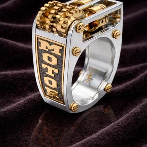 Puede incluir: Un anillo plateado con engranajes y detalles dorados. El anillo presenta la palabra "MOHOR" en letras doradas sobre un fondo negro. Se ven tornillos dorados en el exterior del anillo.