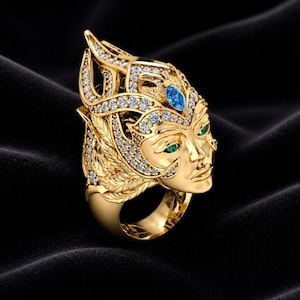 Könnte beinhalten: Ein goldfarbener Ring mit einem detaillierten Gesichtsdesign und Kopfschmuck. Der Ring ist mit klaren und blauen Edelsteinen sowie grünen Edelsteinen für die Augen verziert. Der Ring ist vor einem schwarzen Hintergrund platziert.