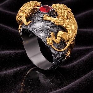 Può includere: Un anello dorato con due figure di leoni dettagliate che fiancheggiano una gemma rossa. I leoni sono posti su una fascia grigio scuro strutturata. Il design dell'anello è intricato e audace, con un tema regale.
