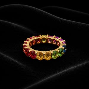 Könnte beinhalten: Ein goldener Ring mit einem durchgehenden Band aus oval geschliffenen Edelsteinen in Regenbogenfarben. Die Edelsteine umfassen rote, orange, gelbe, grüne, blaue und violette Farbtöne. Der Ring ist vor einem schwarzen Hintergrund platziert.