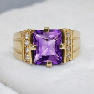 May include: A gold ring featuring a large, square-cut purple gemstone. The ring has a gold band with small, clear gemstones on each side of the central stone. The ring is set against a white background.