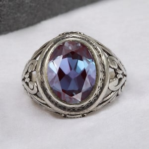 Könnte beinhalten: Silberner Ring mit einem ovalen, facettierten Edelstein in Lila- und Blautönen. Der Ring hat ein verziertes, filigranes Design um den Edelstein und am Band.