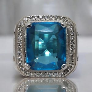May include: A silver ring featuring a large, rectangular, light blue gemstone. The gemstone is surrounded by a halo of smaller, clear stones. The ring has a polished finish and a detailed band.