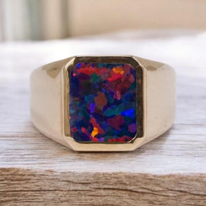 Op de afbeelding: Een goudkleurige ring met een rechthoekige, veelkleurige opaalsteen. De opaal toont een levendige mix van blauw, rood en groen. De ring ligt op een lichtgekleurd houten oppervlak, wat het ontwerp van het sieraad benadrukt. De steen meet ongeveer 1,5 cm x 1 cm.