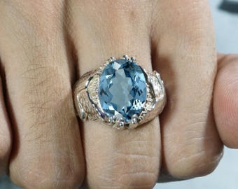 May include: A silver ring featuring a large, oval-cut, light blue gemstone set in the center. The metal band has a textured, organic surface finish that extends from the setting along the sides of the ring, creating a detailed, rugged appearance.
