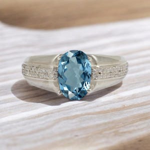 Op de afbeelding: Een zilveren ring met een grote, ovale, lichtblauwe edelsteen. De band is versierd met kleine, heldere stenen. De ring ligt op een lichtgekleurd oppervlak met houtnerf.