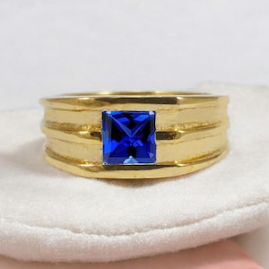 Könnte beinhalten: Ein goldfarbener Ring mit einem quadratischen, tiefblauen Edelstein in der Mitte. Der Ring hat ein dickes Band mit horizontalen Rillen. Der Edelstein ist ein leuchtendes Blau, gefasst in einer goldenen Lünette.