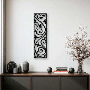 May include: Black metal wall art featuring a vertical rectangular design with an intricate, swirling floral pattern. The artwork is mounted on a white wall, adding a touch of elegance to the space. The design includes leaves and vines.