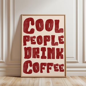 May include: Framed art print with the text "COOL PEOPLE DRINK COFFEE" in red, bold, retro-style lettering. The words are stacked on a cream-colored background, encased in a thin, wooden frame. The artwork is displayed against a white wall.