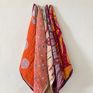 May include: A collection of five folded fabric quilts featuring diverse patterns and colors, including bright orange with white circular motifs, a soft pink with small triangles, a cream base with dark lines, and deep red with floral and leaf designs.