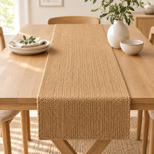 May include: A rectangular table runner made of a thick, tan-colored knit fabric with a prominent vertical ribbed texture. The runner is centered on a light-toned wooden dining table, extending from the front edge toward the center.