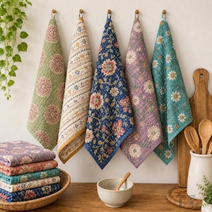 May include: Five quilted kitchen towels with floral and geometric patterns in green, cream, navy blue, purple, and teal hang from hooks. A stack of folded matching towels sits in a woven basket, alongside a ceramic bowl and wooden utensils.