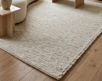 May include: A large, rectangular, off-white area rug with a thick, textured, hand-woven wool construction. The rug features a dense, looped pile surface and a braided edge finish, covering a significant portion of the light-toned wooden floor.