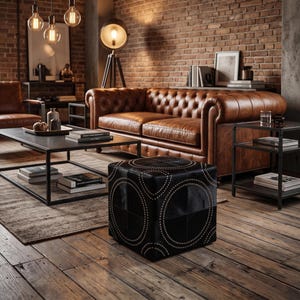 Peut inclure: Un pouf carré noir orné d'un motif géométrique de clous circulaires. Le pouf est placé devant un canapé capitonné en cuir marron, une table basse en métal noir et une table d'appoint en métal noir dans une pièce aux murs en briques.