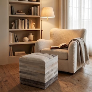Puede incluir: Un sillón texturizado de color crema con una manta a juego sobre el brazo y un libro abierto en el asiento. Delante hay un puf cuadrado construido con tiras horizontales de piel en tonos grises, blancos y marrones.