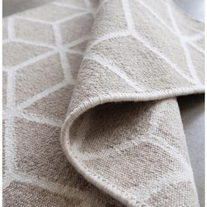 May include: Beige and white geometric patterned rug, partially rolled up. The rug features a repeating pattern of white lines on a beige background. The edge of the rug is visible, showing a stitched border. The rug is made of a textured material.