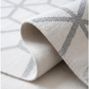 May include: A rolled-up, off-white rug with a geometric pattern in light gray. The rug's edge is visible, showcasing the thick, soft texture and the subtle color contrast between the white and gray design.