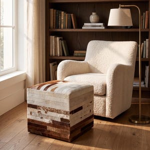 May include: A square ottoman featuring a patchwork of horizontal cowhide strips in shades of white, tan, and dark brown, positioned in front of an off-white textured fabric armchair with rounded arms and a matching floor lamp with a beige shade.