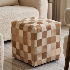 Könnte beinhalten: Ein quadratischer Hocker mit einem Schachbrettmuster aus abwechselnden hellcremefarbenen und mittelbraunen Fellleder-Patches. Der würfelförmige Fußhocker hat eine flache Oberseite und vertikale Seiten, die ein durchgehendes Rasterdesign zeigen.