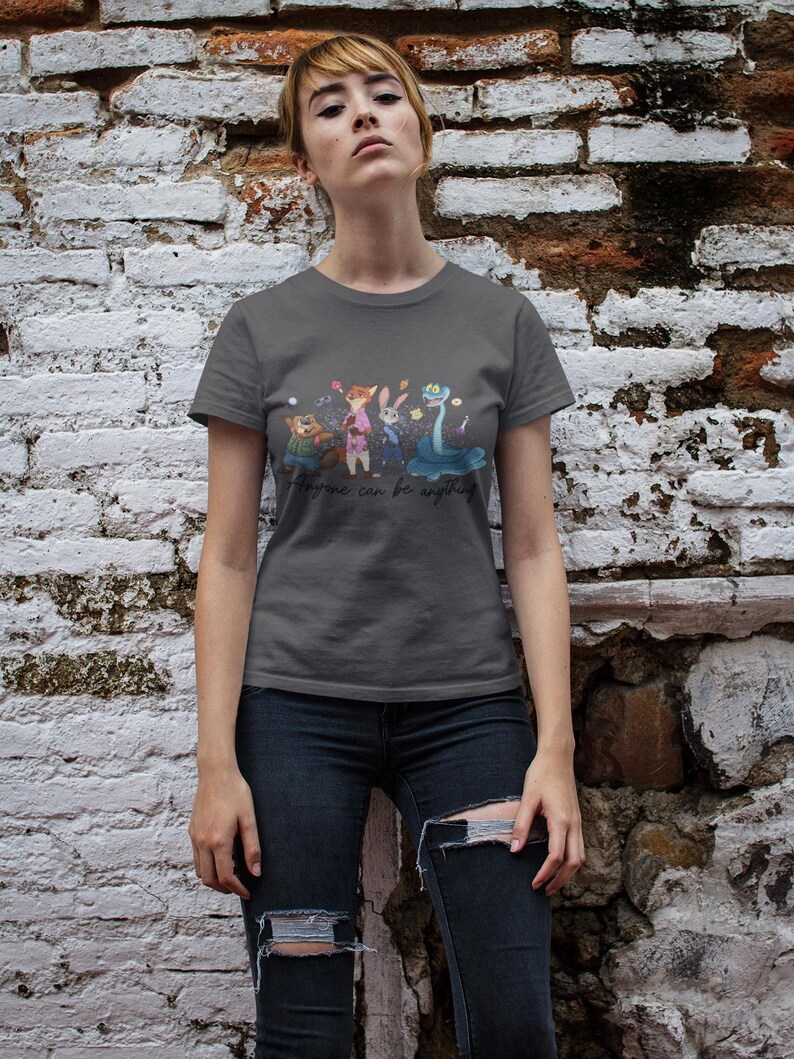 Pu&ograve; includere: Una t-shirt a maniche corte grigio scuro con una stampa grafica di quattro personaggi animati, tra cui un castoro, una volpe, un coniglio e un serpente. Sotto i personaggi, il testo "Anyone can be anything" &egrave; scritto in un carattere corsivo nero.