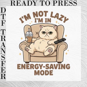 May include: A DTF transfer featuring a cartoon illustration of a beige cat sitting in a beige armchair, holding a drink and a remote control. The text reads READY TO PRESS, I'M NOT LAZY I'M IN ENERGY-SAVING MODE, surrounded by small star accents.