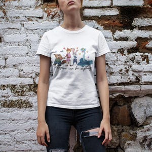 Pu&ograve; includere: Una t-shirt bianca a maniche corte con una stampa grafica di quattro personaggi animati e il testo "Anyone can be anything" in corsivo. I personaggi includono un castoro, una volpe, un coniglio e un serpente blu con piccole stelle.