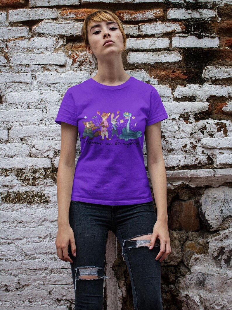 Pu&ograve; includere: Una t-shirt viola a maniche corte con una stampa grafica di quattro personaggi animati, tra cui una volpe, un coniglio e un serpente, con il testo "Anyone can be anything" stampato in corsivo nero al centro della zona del petto.