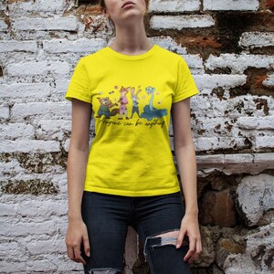 Pu&ograve; includere: Una t-shirt a maniche corte di colore giallo brillante con una stampa grafica di quattro personaggi animali animati e il testo in corsivo "Anyone can be anything" stampato in nero al centro del petto.