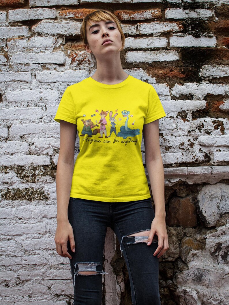 Pu&ograve; includere: Una t-shirt a maniche corte di colore giallo brillante con una stampa grafica di quattro personaggi animali animati e il testo in corsivo "Anyone can be anything" stampato in nero al centro del petto.