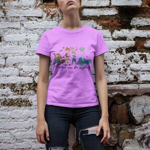 Pu&ograve; includere: Una t-shirt a maniche corte color viola chiaro con una stampa grafica di quattro personaggi animali animati e il testo "Anyone can be anything" in un carattere corsivo nero. La maglietta ha un girocollo e una vestibilit&agrave; standard, indossata con jeans scuri.