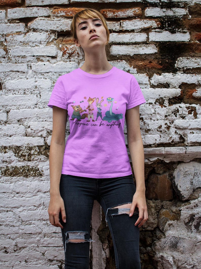 Pu&ograve; includere: Una t-shirt a maniche corte color viola chiaro con una stampa grafica di quattro personaggi animali animati e il testo "Anyone can be anything" in un carattere corsivo nero. La maglietta ha un girocollo e una vestibilit&agrave; standard, indossata con jeans scuri.