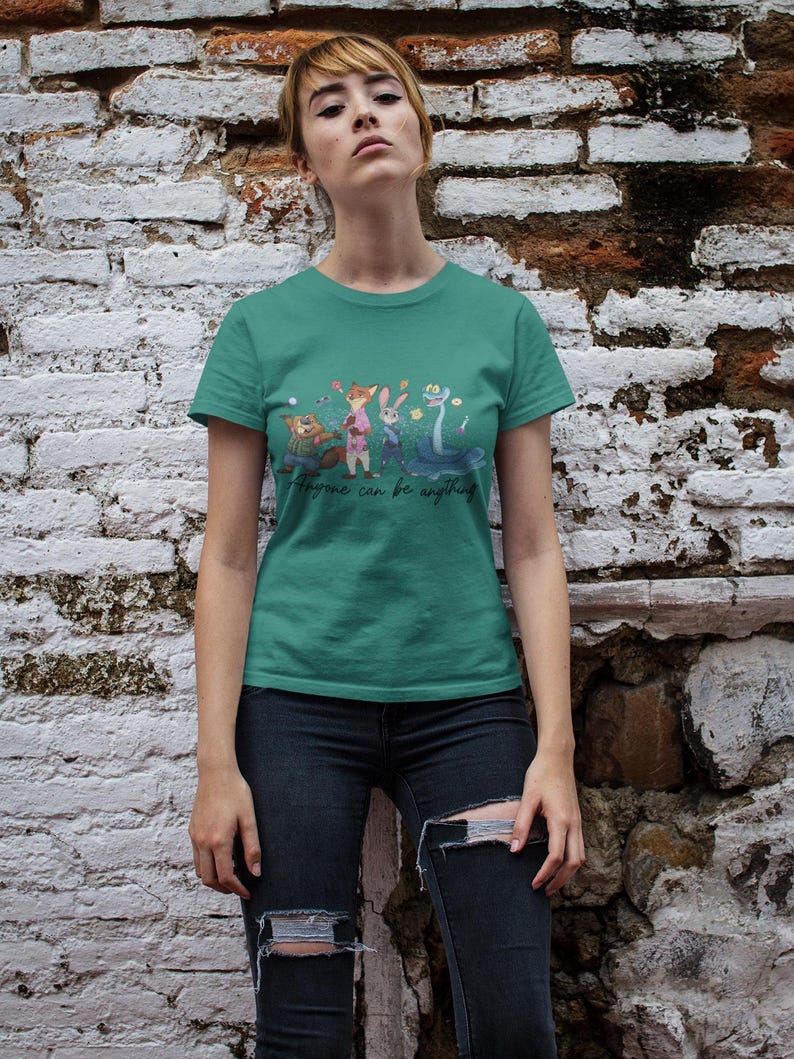 Pu&ograve; includere: Una t-shirt a maniche corte color verde acqua con una stampa grafica di quattro personaggi animati e il testo "Anyone can be anything" in corsivo. La maglietta presenta un girocollo e un orlo dritto con un'illustrazione colorata.