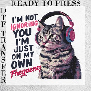 May include: A DTF transfer featuring a gray tabby cat wearing pink and blue headphones. The text reads: READY TO PRESS, I'M NOT IGNORING YOU I'M JUST ON MY OWN Frequency. The design uses dark blue, pink, and gray tones on a white background.